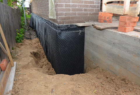 The image shows a construction site where a foundation wall is being waterproofed. The foundation is exposed, and a black dimpled waterproof membrane is applied along the exterior surface. The membrane helps prevent water infiltration into the building's foundation. The area around the foundation has been excavated to allow for the installation of the membrane. The exposed ground and soil are visible alongside the wall, and a temporary wooden structure is present on the right side, possibly for construction support. A wooden fence runs along the left side of the area.