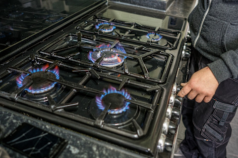 An individual is standing beside a gas stove, with their hand resting on the controls. The burners are ignited, showing a clear blue flame, indicating the stove is on and functioning. The focus is on the stove's surface, reflecting a careful check or adjustment being made, possibly during a safety inspection or routine maintenance.