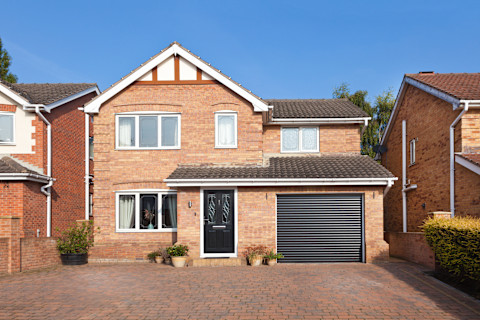 This image shows a modern brick house with two stories. The house has a dark-colored front door and a large window on the left side. On the right, there is a garage with a dark gray roller shutter door. The house is surrounded by neighboring properties, which have a similar design. The driveway is paved with light brown bricks, and potted plants are placed near the entrance, giving a welcoming touch. The sky is clear and blue, providing a bright, sunny backdrop for the scene.