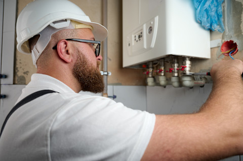 A male boiler engineer wearing protective equipment and glasses examines a newly installed gas boiler with four pipes on the bottom, going into the tiled wall. In his hand is a screwdriver, which is pressed against one of the boiler pipes.