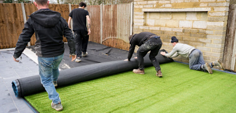 This image shows four landscapers working together to unroll artificial grass in a back garden.