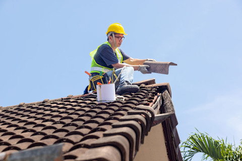 Worker repairing leaking ridge tiles on a tiled roof under a clear blue sky.

