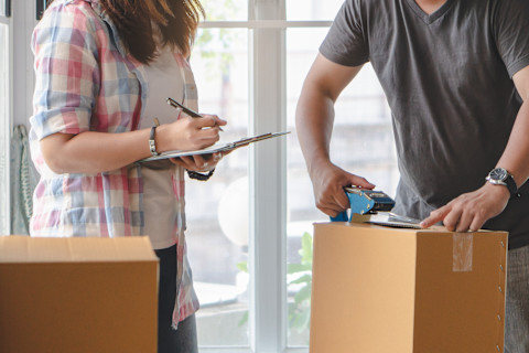 This image shows a couple packing a cardboard box. A woman is checking things off on a clipboard ensuring nothing is forgotten. 