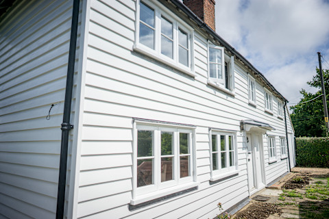 A two-story, traditional white clapboard house with black trim around white-framed windows, a white door, and a downspout on the left side, under a blue sky with clouds.