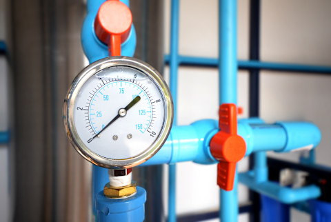 Close-up of a water pressure regulator installed on a plumbing system. The image shows the regulator with a gauge, connected to pipes. The background includes other plumbing components and fixtures.