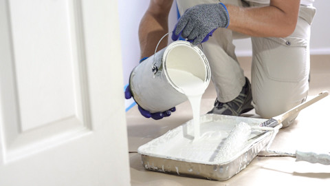 This image shows a painter wearing overalls pouring a tin of white paint into a tray with a roller resting on the side.