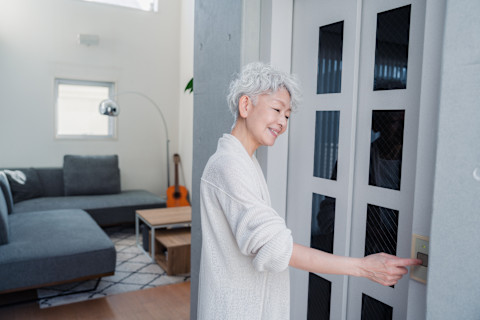 In a living room with white walls, gray sofas, and a guitar in the background, a woman presses a button to operate her modern stair lift. The lift, which is sleek and white with large glass windows, is ready to take her up the stairs.