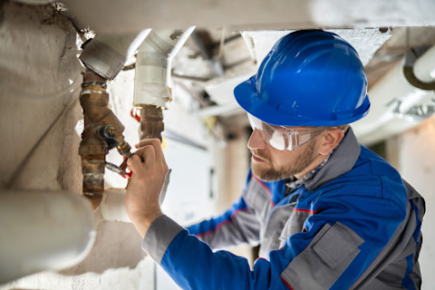 Close-up of a technician adjusting a water pressure regulator. The image shows the technician working on a valve or gauge, with plumbing tools and equipment visible. The background includes pipes and fixtures related to the water system.