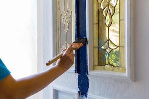 This image shows a close-up of a person using a paintbrush to apply vibrant blue paint to an exterior front door.