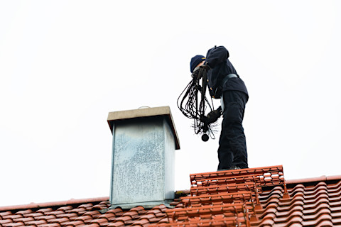 This image shows a chimney sweep standing on a residential rooftop with equipment, preparing to clean the chimney.