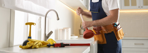 A person using a plunger to unblock a kitchen drain, with a sink full of water and cleaning supplies nearby.