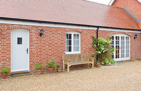 House extension providing a single storey granny annexe. It is a red brick building with a white entry door, patio doors, and window. Beneath the window is a wooden bench on a gravel lawn. Several potted plants and a climbing plant are outside.