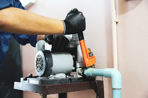 A man dressed in a blue jacket and black gloves is holding an orange tool while working on a pale blue water pump. He is adjusting something at the end of the pump.