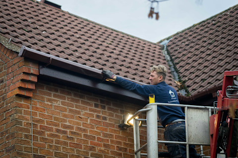 A professional installing gutters along the edge of a roof, using a ladder and tools, with newly installed gutters being fitted to the roofline.