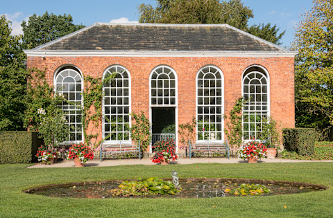 This image shows a red-brick orangery in a manicured garden. There is a circular pond in the foreground and potted plants near the front walls of the orangery. The orangery has traditional grid windows.