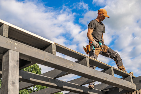 A skilled construction worker is standing on a wooden framework, carefully placing roof panels.
