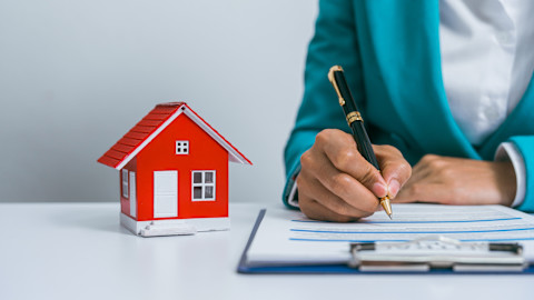 This image shows a professional sitting at a desk signing a contract next to a small model of a house.