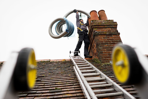 A worker standing on a roof near a chimney stack, carrying a large coiled hose.