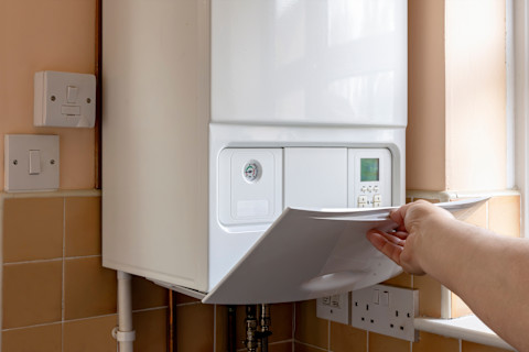 A hand holds open the control panel on a gas boiler, situated on a tile wall in a kitchen. The boiler's controls are visible under the panel.