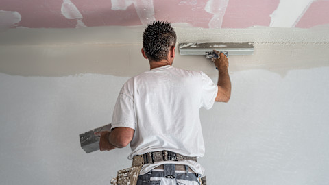 This image shows a tradesperson on a ladder skimming the top of a wall, close to the ceiling. He is using special stainless steel taping knife.