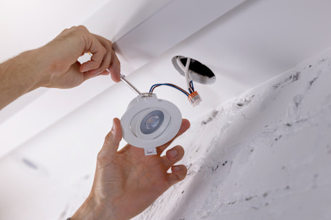 A professional installing recessed lighting in a ceiling, with the ceiling open to reveal the light fixtures being positioned and secured, and tools and materials visible nearby.