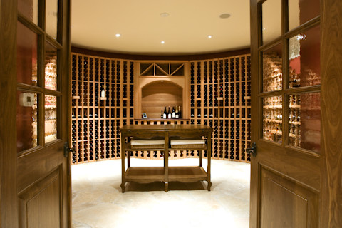 A large, sparsely stocked home wine cellar room with wooden storage shelves and a wooden table in the centre