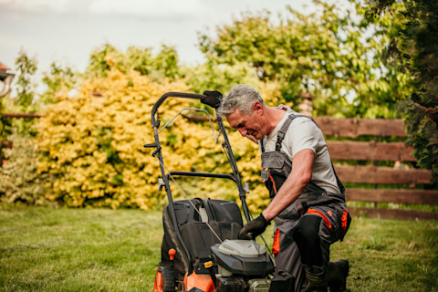A middle-aged man is kneeling on the grass in a backyard while working on a lawn mower. He is wearing work overalls and gloves, indicating that he is performing maintenance or repair on the machine. The setting is outdoors with a garden in the background, featuring green trees and yellow bushes. The man appears focused on his task, and the image conveys a sense of hands-on repair work.