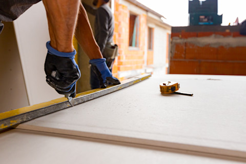 A close-up image of a worker cutting a sheet of plasterboard with a precision utility knife. The worker’s hands are firmly holding the knife and guiding a metal straightedge ruler to ensure a clean, straight cut. The plasterboard is laid flat on a work surface, and the background features construction materials and tools, indicating an active building or renovation site.