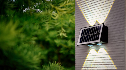 A motion sensor, solar-powered wall light emits bright cross-shaped beams, mounted on a white, textured wall. Lush green foliage, blurred in the background and foreground, creates a fresh, eco-friendly feel.