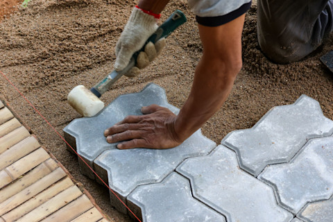 This image shows a close-up of interlocking concrete paver tiles laid in a modern, uniform pattern on a compacted sand base. The tradesperson carrying out the tasks is using string as a guide and a mallet to ensure the blocks are fitted correctly.