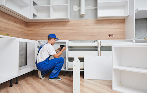 A worker in blue overalls uses a drill to assemble white kitchen cabinets on brown wood flooring. The setting is a partially constructed kitchen with a focused atmosphere.