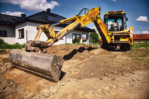 An excavator working on a house construction site, digging the ground with a bucket attachment. The machine is surrounded by construction materials, dirt, and an ongoing construction project.