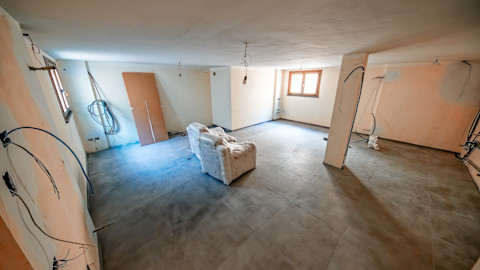 A large, vacant basement room with two solitary chairs in the centre and a hanging light bulb overhead. On the walls around the room are several exposed wires ready for electrical connections. Two high windows on either side of the room let in bright sunlight.