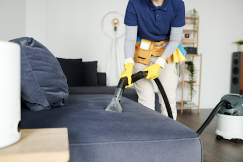 A professional cleaning service worker wearing yellow rubber gloves and a tool belt uses a vacuum cleaning device to remove dust and dirt from a blue-grey fabric sofa in a well-lit modern living room.