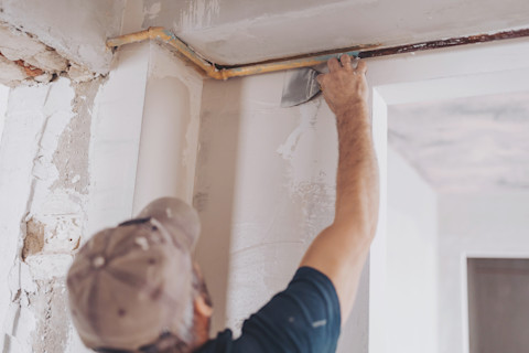 A male plasterer wearing a baseball cap reaches up to the top of a wall of a house under renovation to spread plaster using a finishing tool.