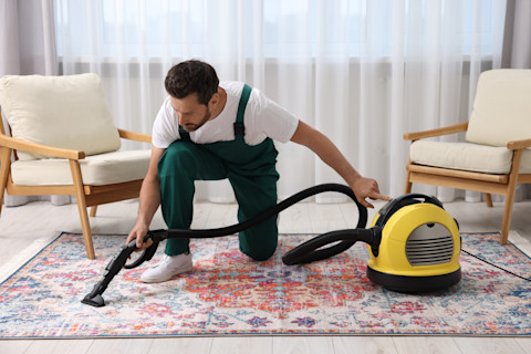 A professional carpet cleaner using a steam cleaning machine on a beige carpet in a living room, with furniture carefully moved aside.