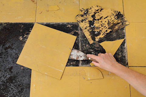 A hand uses a scraper to remove damaged yellow linoleum tiles from a floor during a renovation project, revealing blackened adhesive underneath.