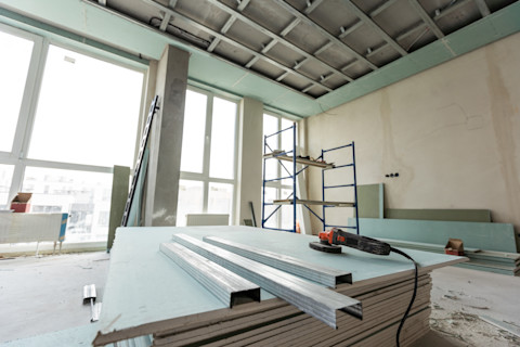 The working process of installing metal frames for plasterboard in a shop under renovation. A stack of plasterboard in the centre has three metal frames and an electrical grinder on top, with a metal shelfing unit in the background against three large windows.