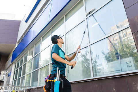 A window cleaner using a squeegee and cleaning solution on a large glass window, with a clear view of the outdoors and a ladder positioned nearby.