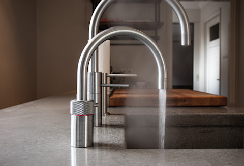 This image shows a quooker tap, a modern boiling water tap in a sleek and modern kitchen. Water is running from the tap and there is the evidence of steam suggesting the water running from the tap is of a very high temperature.