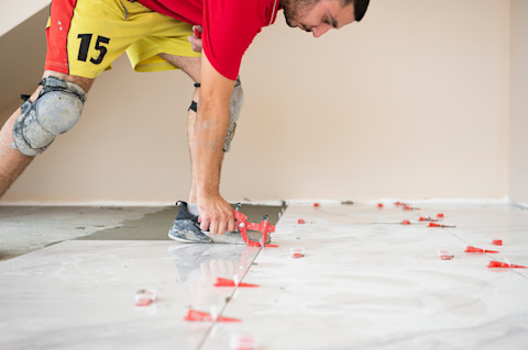 Man in red shirt and yellow shorts installing floor tiles with spacers and a levelling tool in a domestic bathroom, a look of precision and focus on his face. 