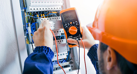 This image shows an electrician wearing an orange hardhat using a digital tool to check the wiring in a circuit breaker is functioning correctly. 