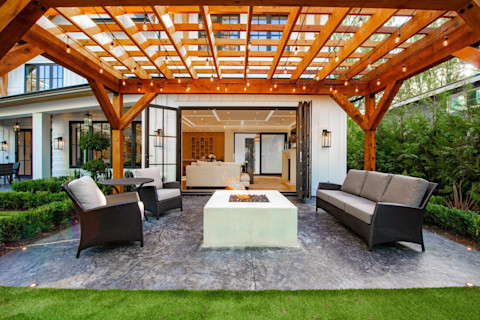 This image shows a serene backyard scene featuring lush green grass, a stone fire pit surrounded by comfortable seating and a backdrop of plants and trees. The warm sunshine creates a peaceful atmosphere, highlighting the beautiful landscaping.