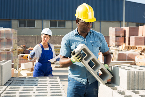 Close-up of a worker installing an air brick into the wall of a house. The worker is placing the perforated brick into a prepared opening, with tools and construction materials visible nearby. The surrounding brickwork and mortar are clearly seen.