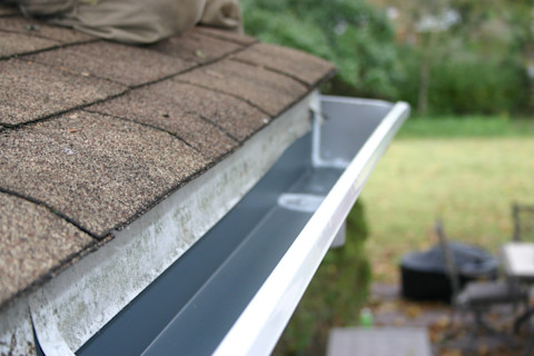 A close-up of a clean, freshly installed house gutter along a roof with brown shingles.