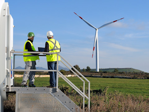 The image shows two workers standing on a metal platform at the base of a wind turbine. They are both wearing high-visibility safety vests and hard hats, one green and one white, and are looking at a laptop screen, possibly reviewing data or plans. In the background, a large wind turbine with red-tipped blades stands in a grassy field under a clear blue sky. The landscape includes rolling hills and sparse vegetation, indicating a rural setting, likely part of a wind farm. The scene conveys a sense of renewable energy operations and maintenance work.
