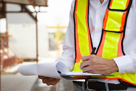 A person in a white shirt and neon safety vest is writing on a clipboard. They're at a construction site, which is blurred in the background, focusing on their work.