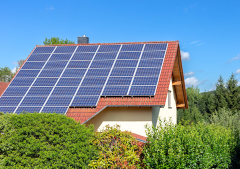 This image shows a number of solar panels on the roof of a house. The roof is made from red/orange tiles and it contrasts against the off-white exterior of the home. There are full, green bushes in the foreground obscuring the bottom half of the property. There is a blue sky and some white clouds.