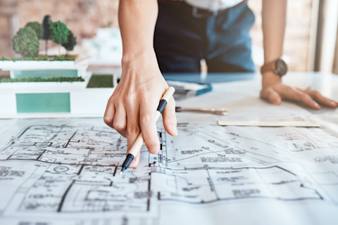 This image shows an architectural professional pointing at architectural drawings on a large piece of paper with their right hand. They are pointing with their middle finger and they have a pen between their forefinger, thumb and middle finger. There is also a white ruler resting on the paper.