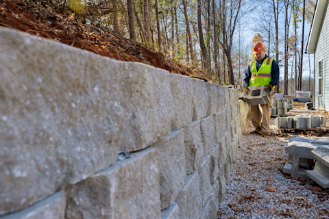 A large, stone garden wall under construction. Already half built, the wall stands 3 stones deep, and a construction worker in a hi-vis and a hard hat can be seen at the far end of the wall adding a new brick to the wall.